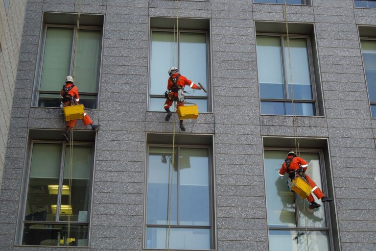Three men in orange safety gear are suspended from a building, performing window cleaning services.